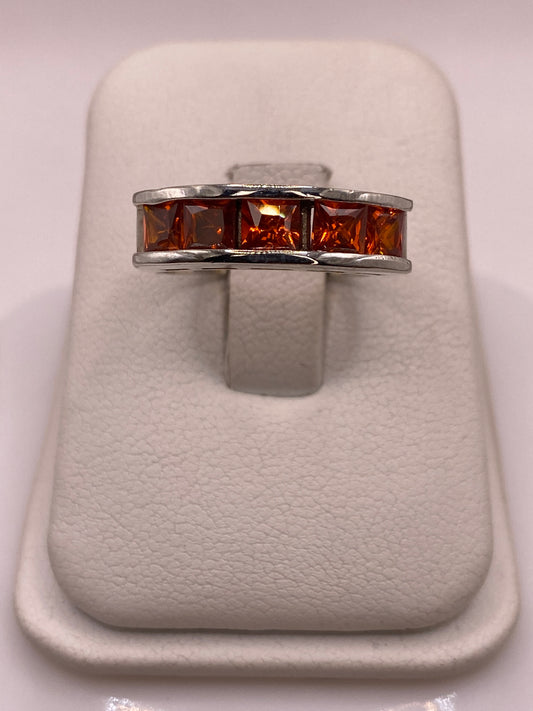 Anillo banda de plata .925 con cinco circonias naranja ámbar de corte princesa en engaste de canal, vista frontal sobre soporte blanco