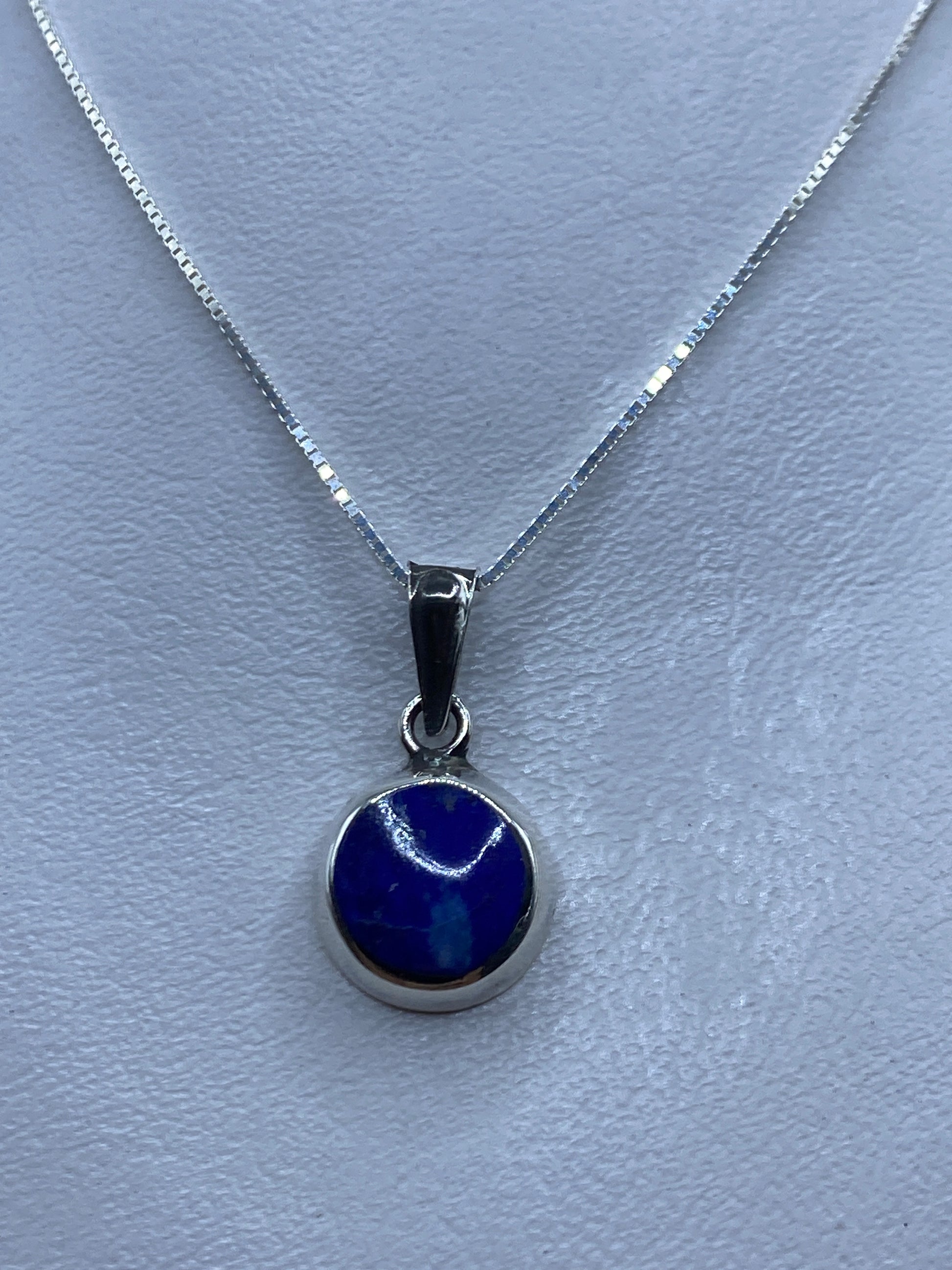 Dije de plata .925 con piedra azul engastada, colgando de cadena de plata sobre fondo azul