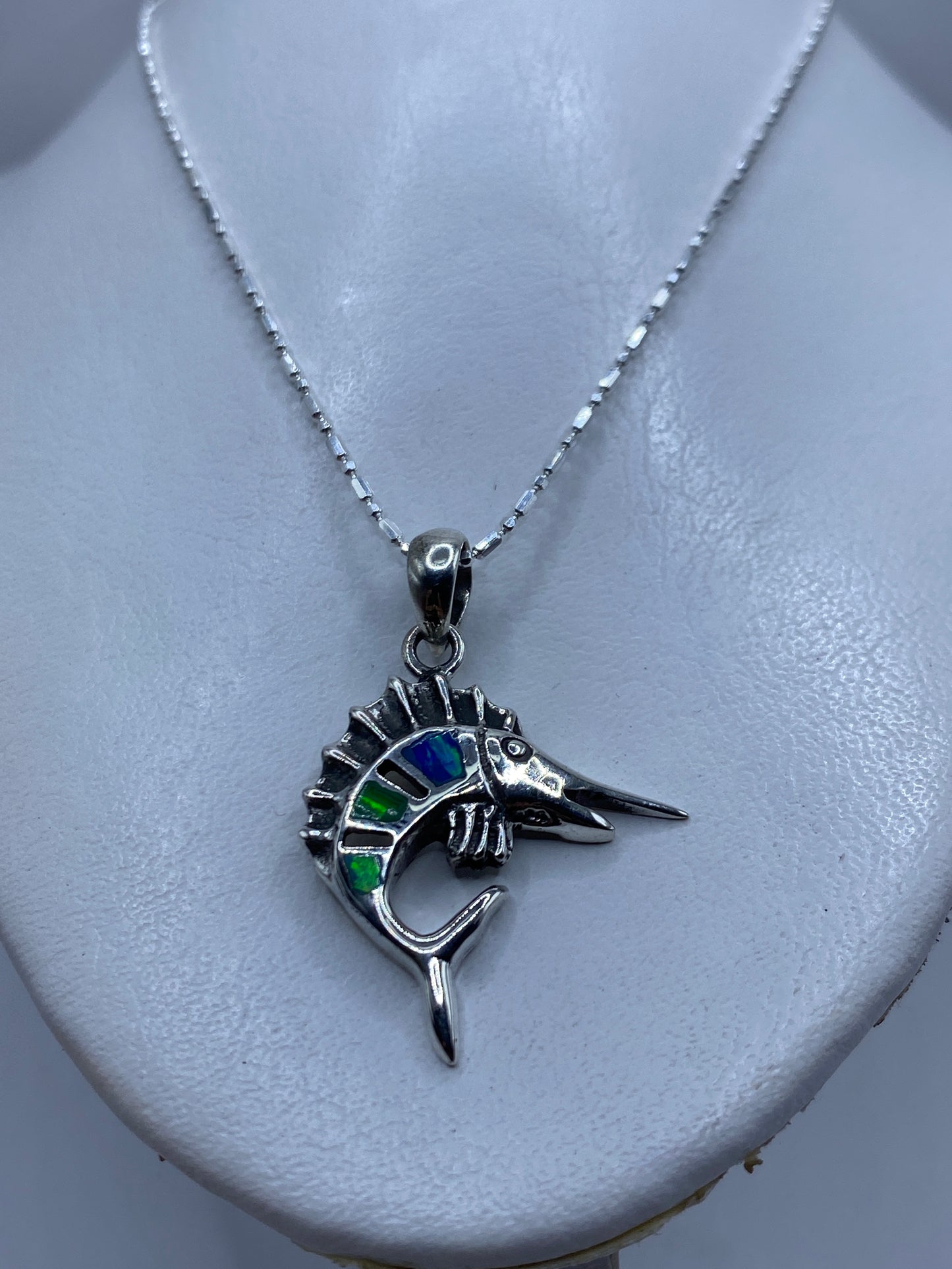 Detalle frontal del dije pez espada con ópalo de fuego reflejando tonos azules y verdes
