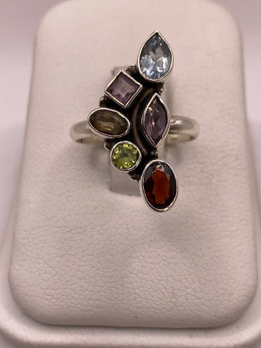 Anillo de plata .925 con seis piedras multicolor — azul, morado, verde y granate — en cortes variados, visto de frente sobre exhibidor blanco