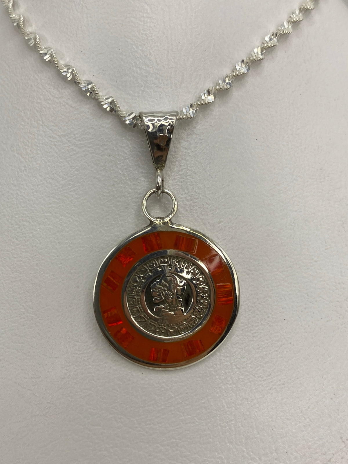 Vista frontal del dije de plata: moneda prehispánica rodeada de ópalo naranja