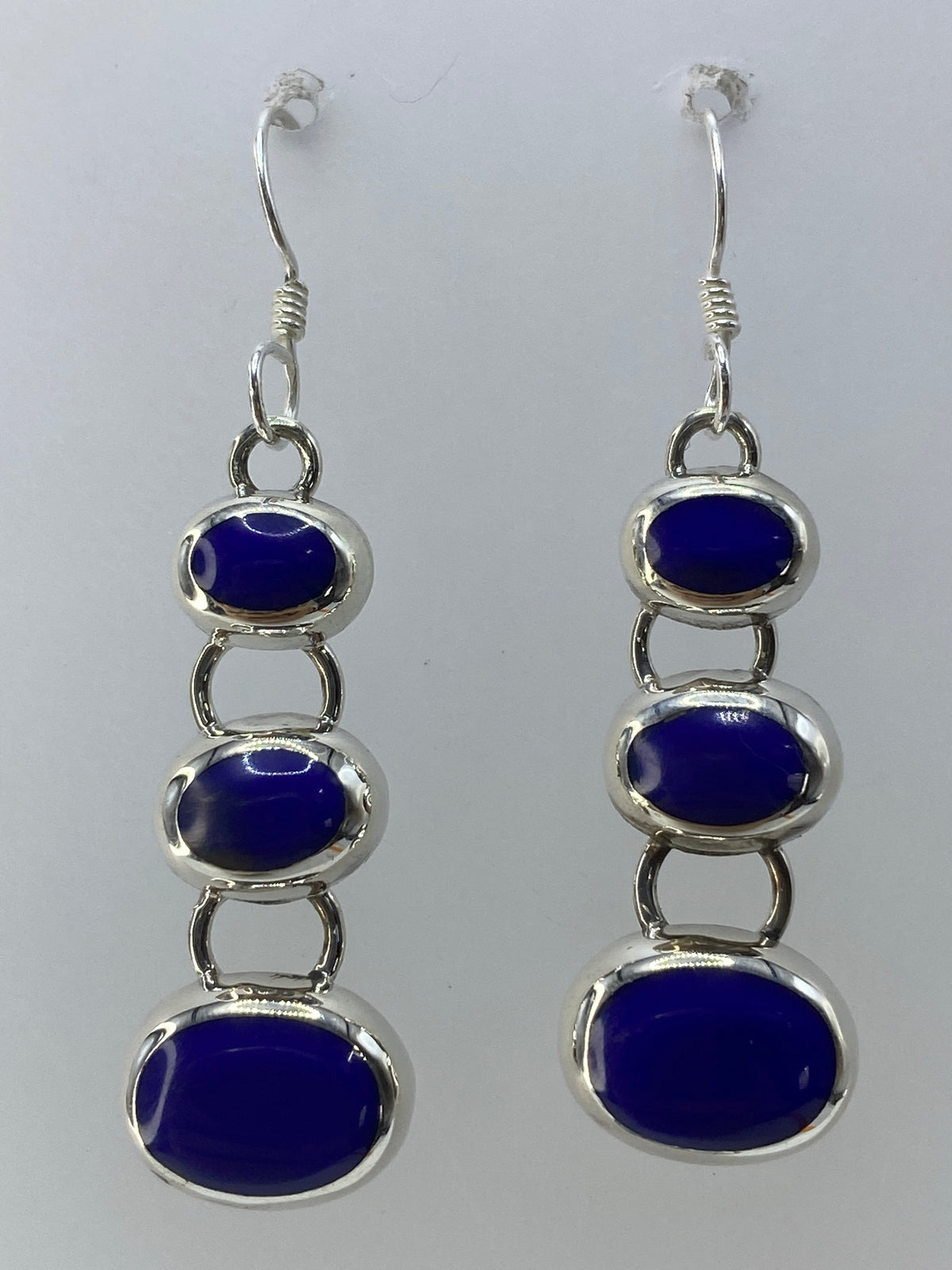Par de aretes de plata con piedra azul en cascada de tres óvalos, colgando sobre fondo blanco