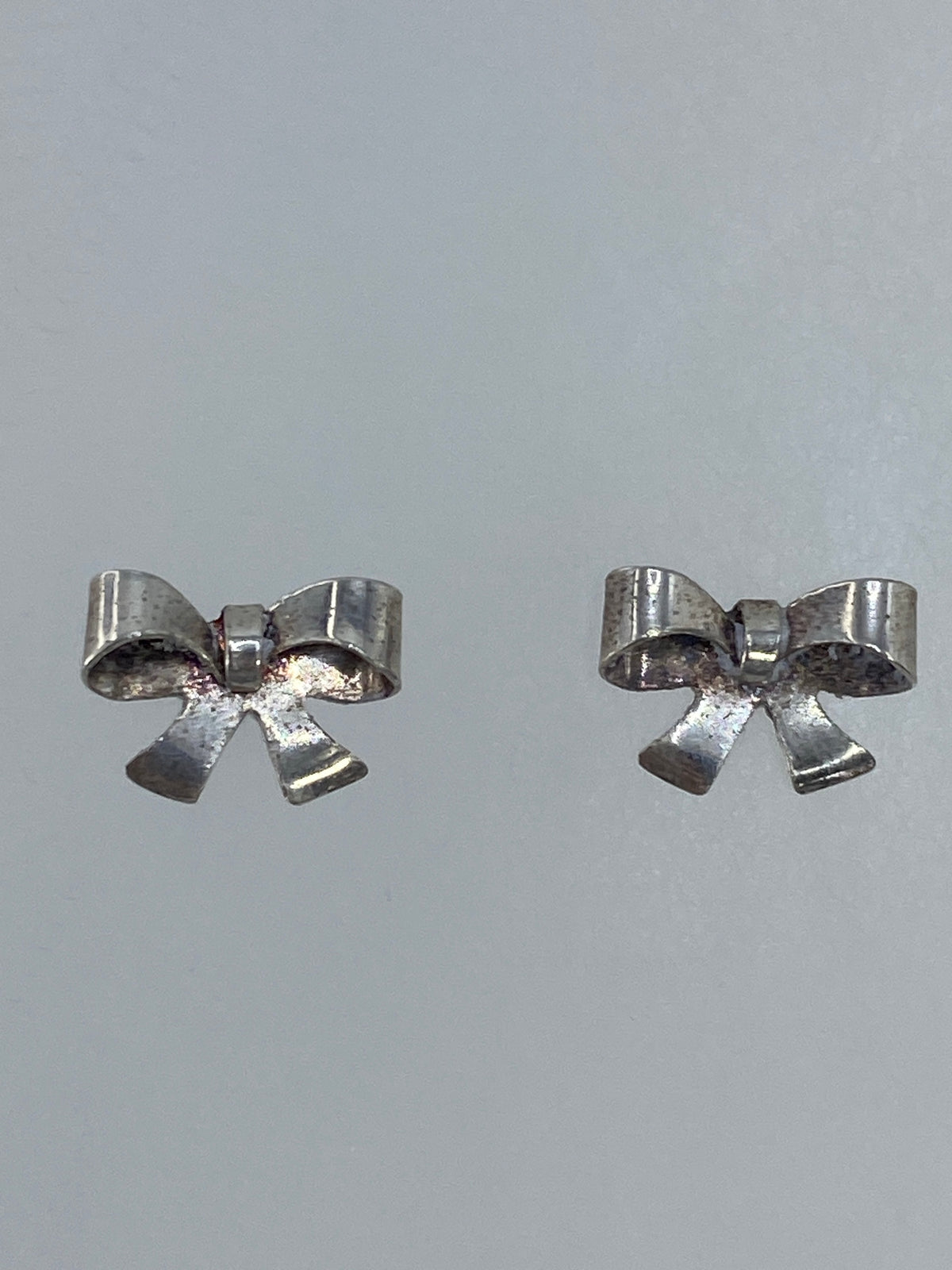 Par de aretes de plata en forma de moño minimalista, 1 cm de largo, sobre fondo gris