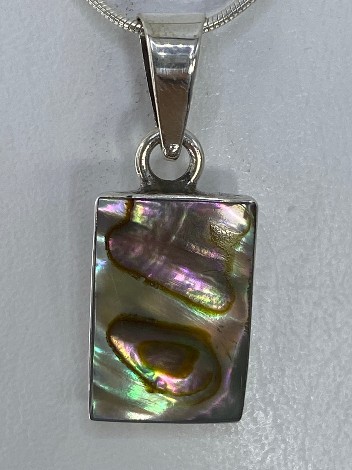Vista frontal del dije rectangular con piedra multicolor que refleja tonos púrpura, verde y dorado