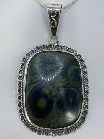 Dije de plata 950 con piedra verde incrustada, colgando de una cadena de plata frente a fondo blanco