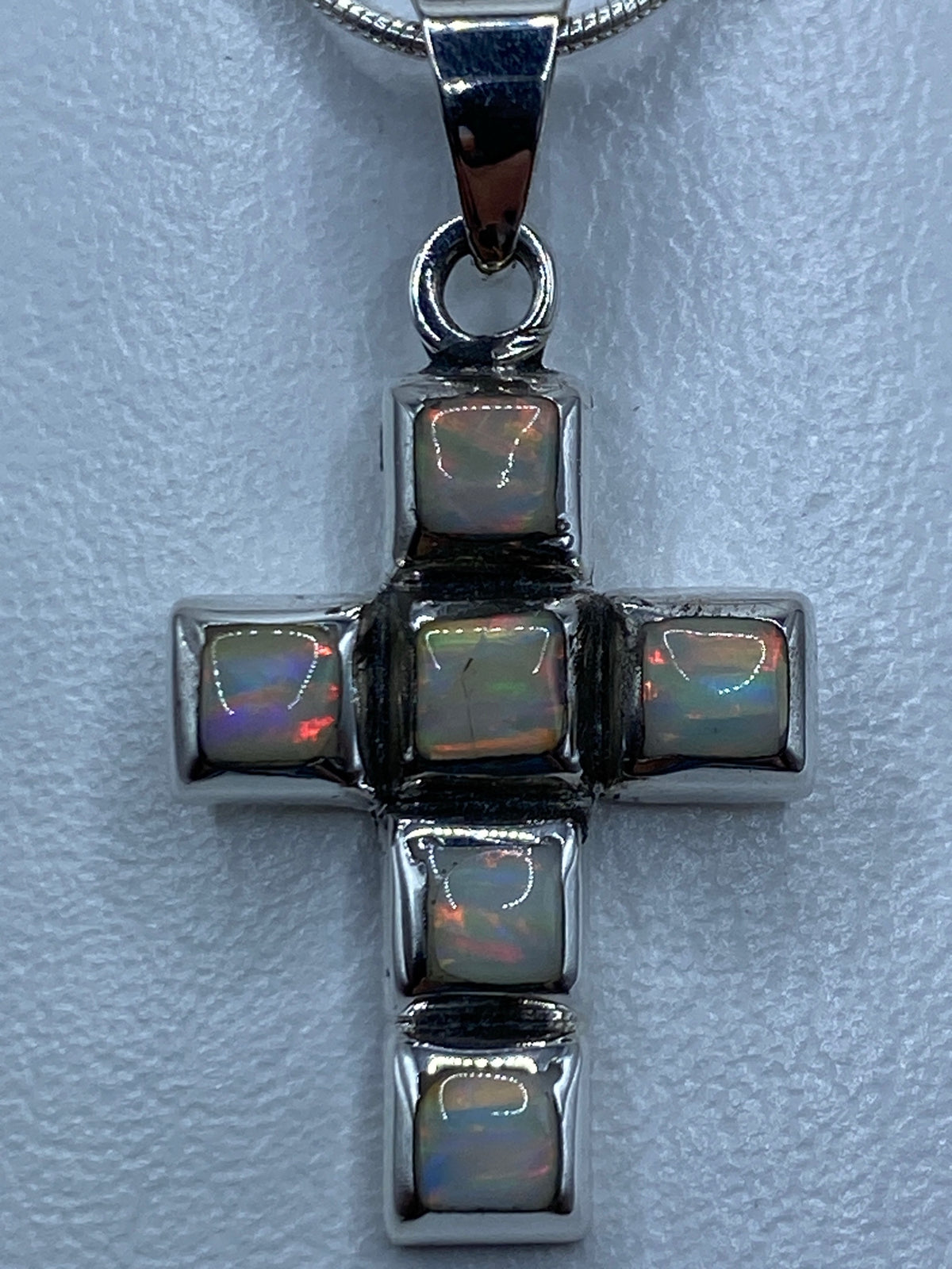 Vista frontal del dije cruz: plata pulida con piedra de color tornasol en el centro