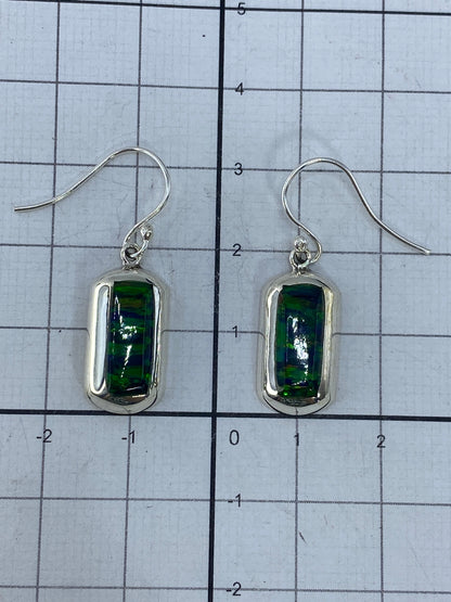 Aretes de plata colgando, mostrando el engaste a mano del ópalo y la refracción de luz verde