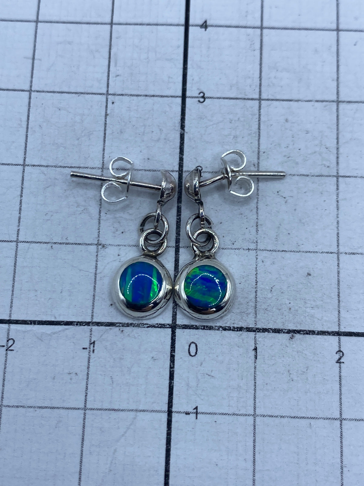 Vista frontal de los aretes con piedra de ópalo en engaste de plata, juntos sobre fondo plateado