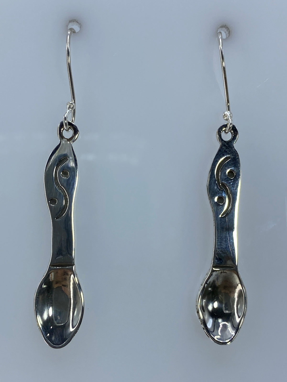 Par de aretes de plata repujados en forma de cuchara, colgando sobre fondo gris