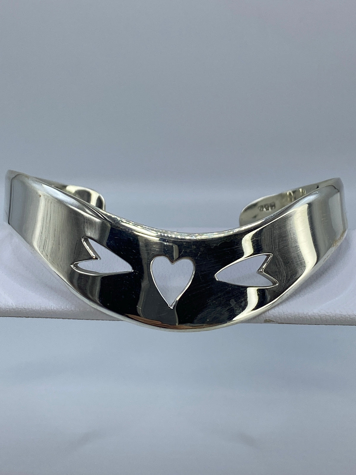 Brazalete rígido de plata en calado con motivo de corazón y alas, vista frontal sobre fondo gris