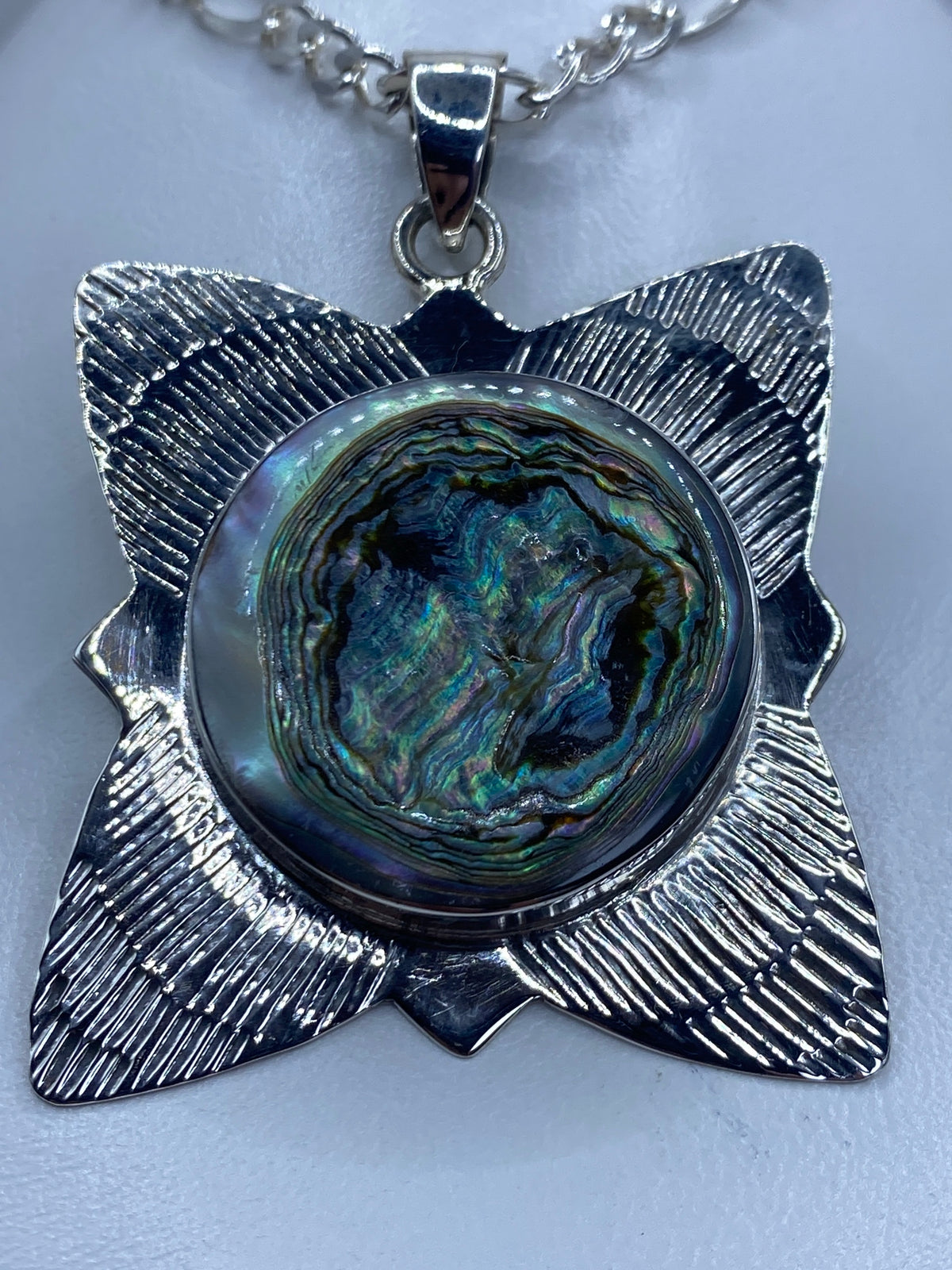 Dije de plata con concha de abulón iridiscente en forma de flor de cuatro pétalos, colgando de una cadena plateada