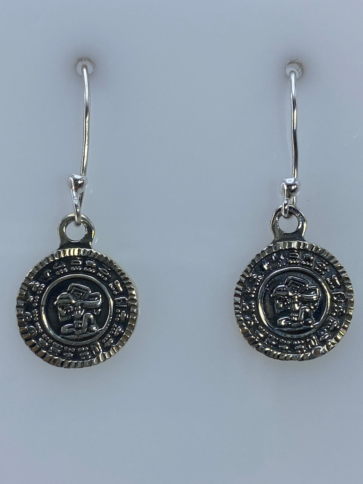 Par de aretes de plata 950 con motivo prehispánico repujado, vistos de frente sobre fondo gris