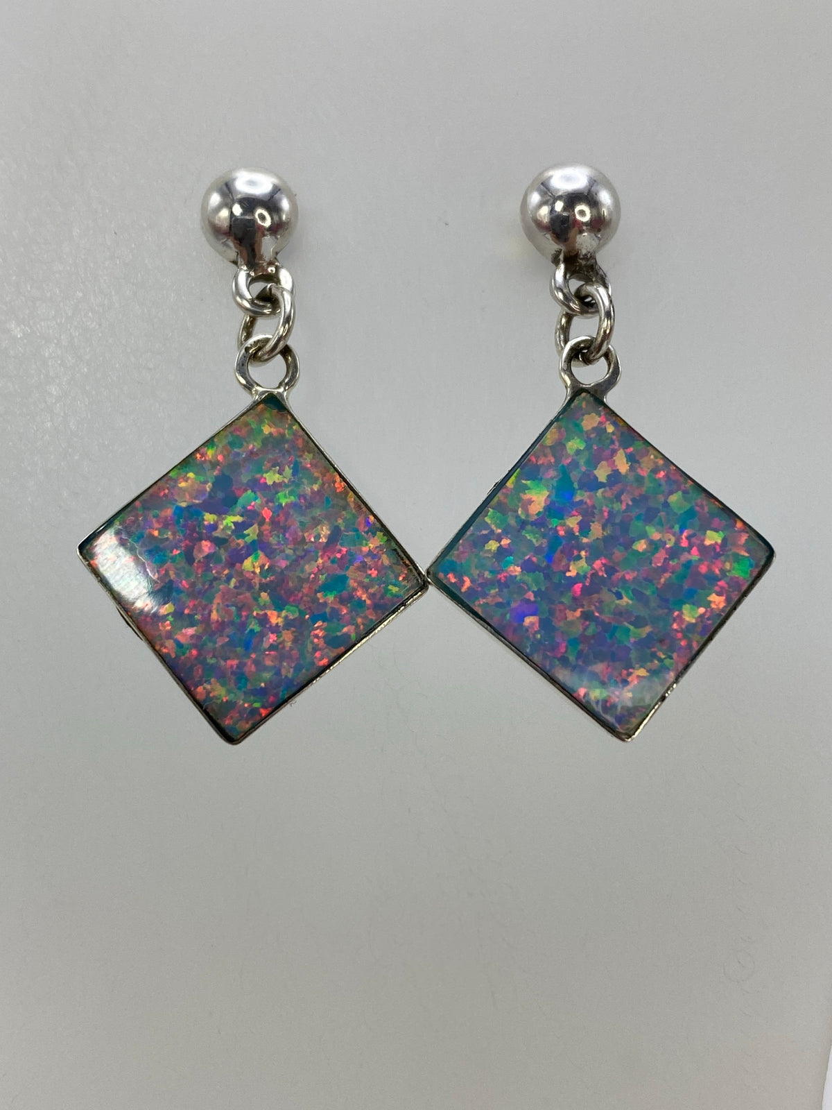 Aretes de plata con ópalo de fuego multicolor colgando sobre fondo blanco, mostrando los destellos de color