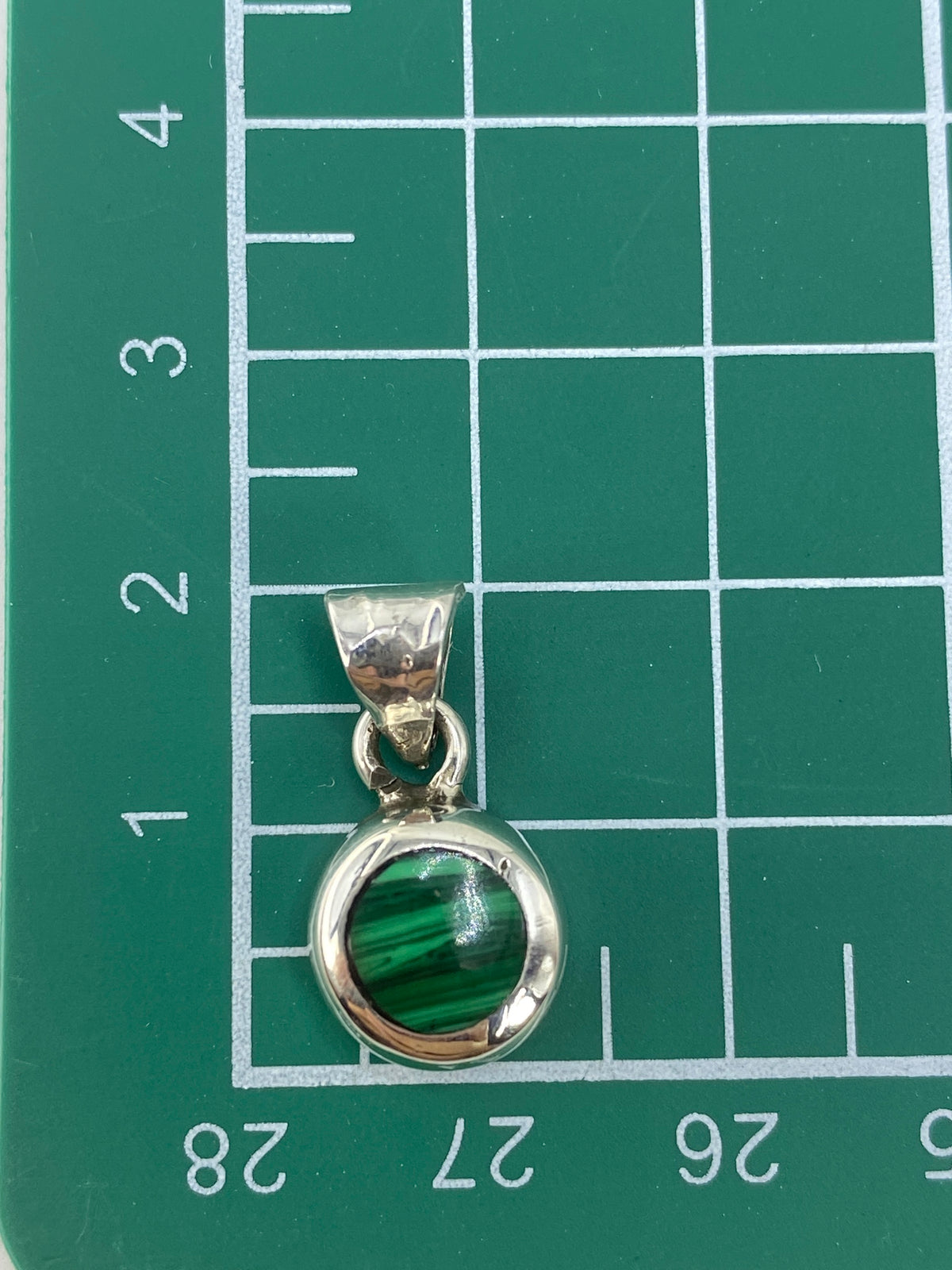 Dije de plata verde sobre regla de medidas confirmando tamaño de 2 cm