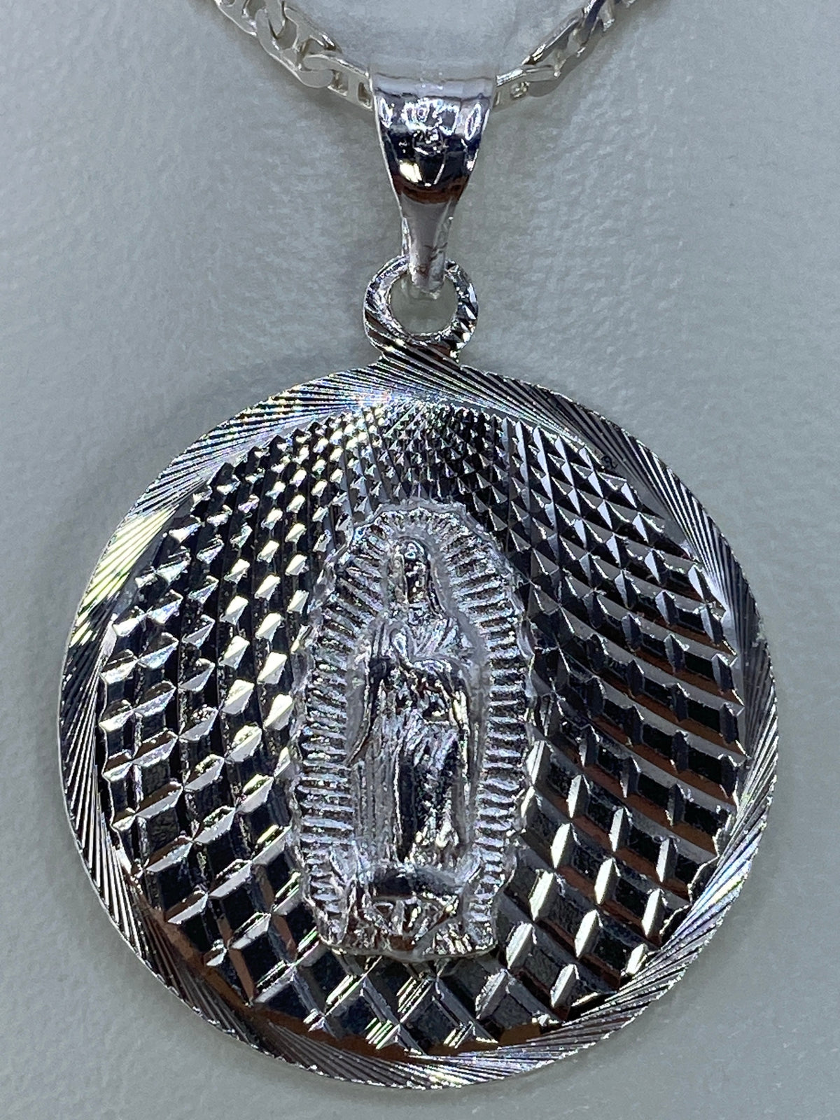 Dije oval de plata sobre fondo blanco, vista frontal completa del motivo religioso