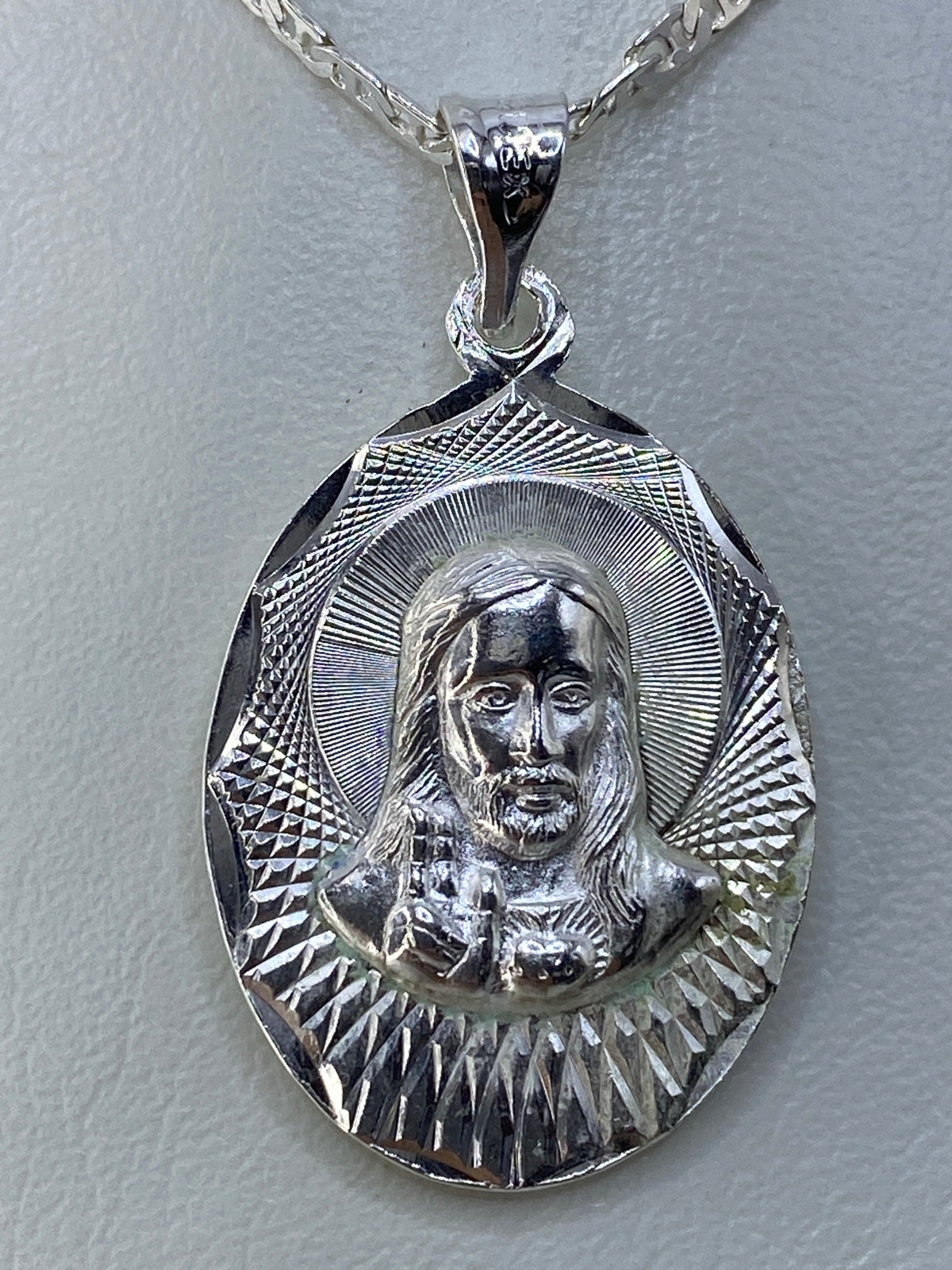 Dije de plata repujado con motivo religioso de 3.5 cm, colgando de cadena plateada