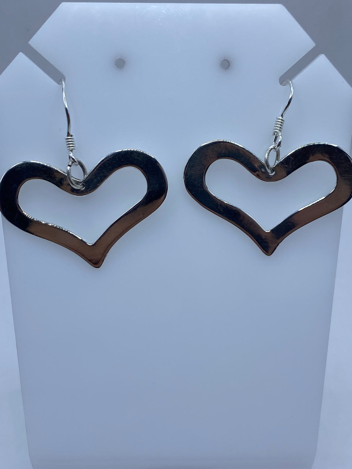 Par de aretes de plata con forma de corazón calado, colgando de ganchos de plata en pieza de exhibición blanca